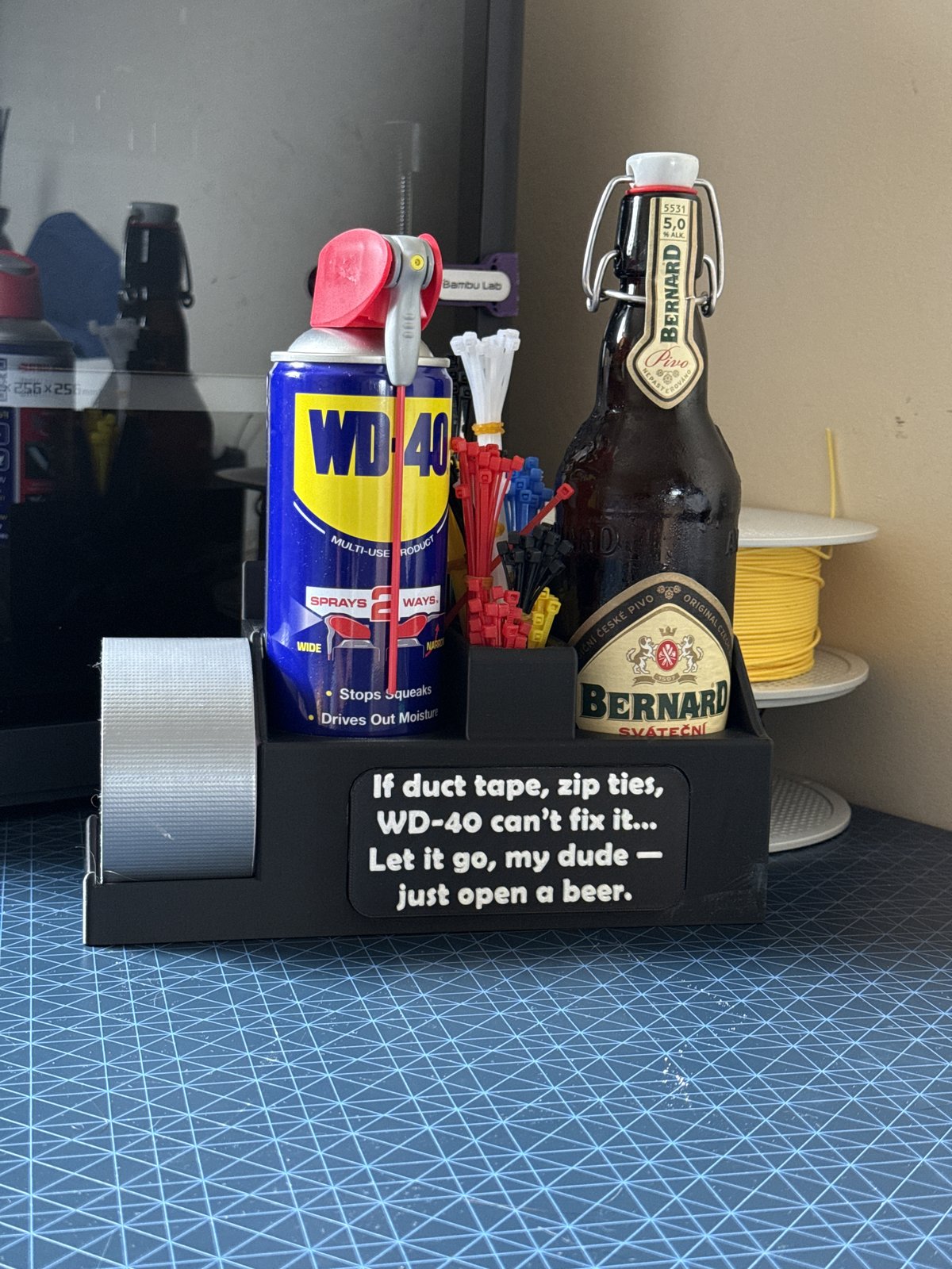 A black 3D-printed mechanic's caddy holding a can of WD-40, coloured zip ties, a roll of duct tape, and a bottle of Bernard beer, with an engraved plaque on the front