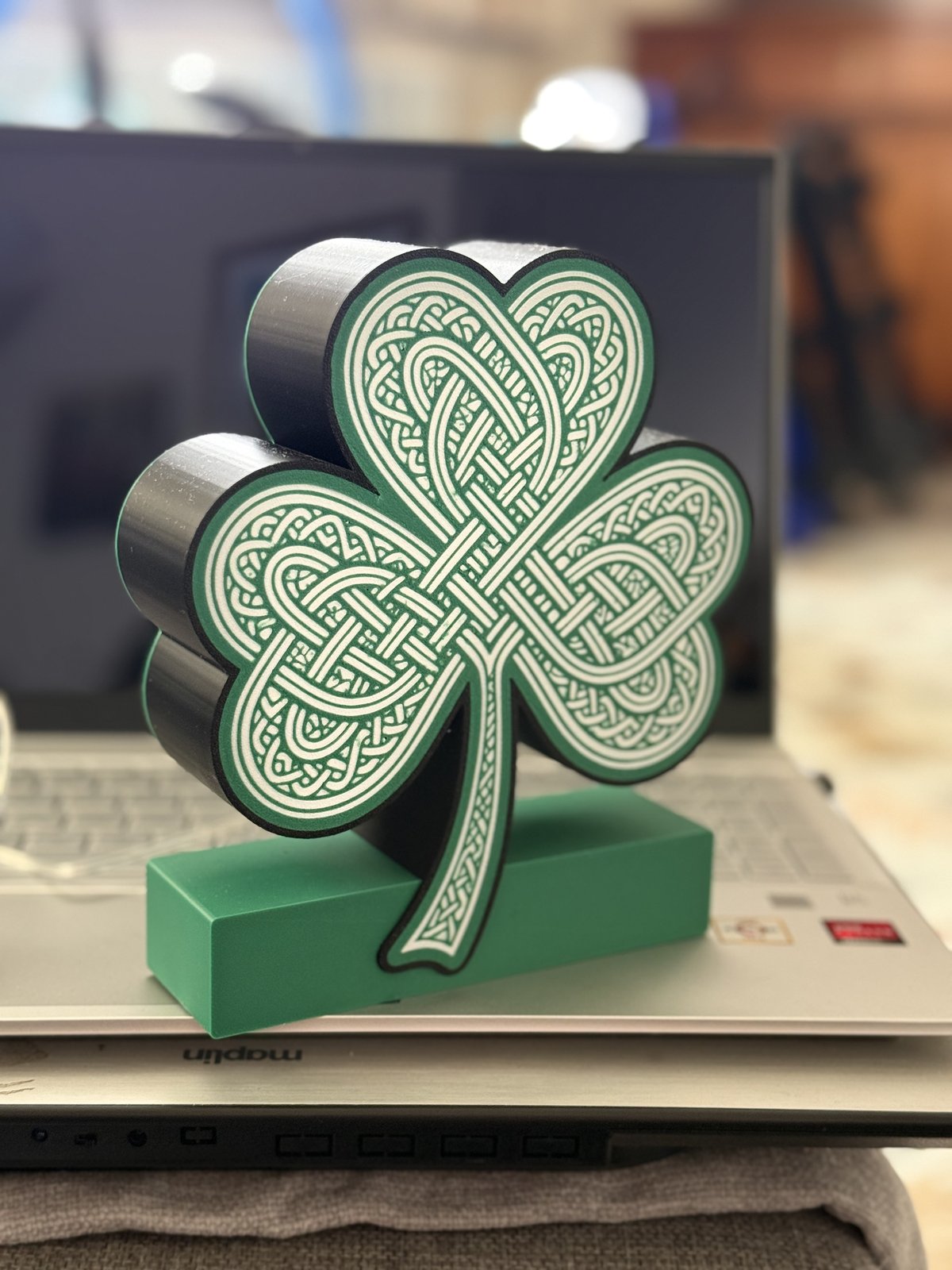 The same Celtic knotwork shamrock LED piece unlit, standing on a laptop in daylight, showing the printed detail